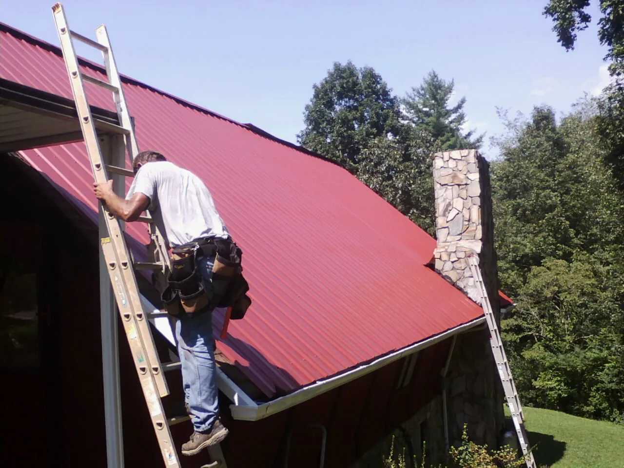 Roofer on a ladder beside a red metal roof and stone chimney — Soffit & Fascia Repair by Roofing Brothers in Rockledge